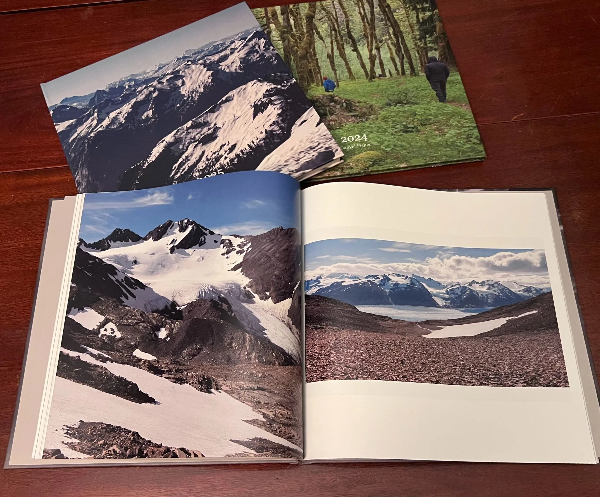 Three printed hardcover photo books on a wooden table. Two closed books, with covers of snowy mountains and a mossy forest marked 2024, lie behind an open book showing a glacier on the left page and a panorama of mountains above an icefield on the right.